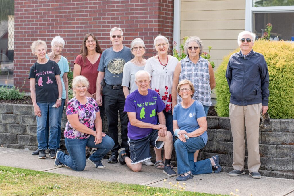 Group photo of older adults and volunteers standing and kneeling outside a brick building.