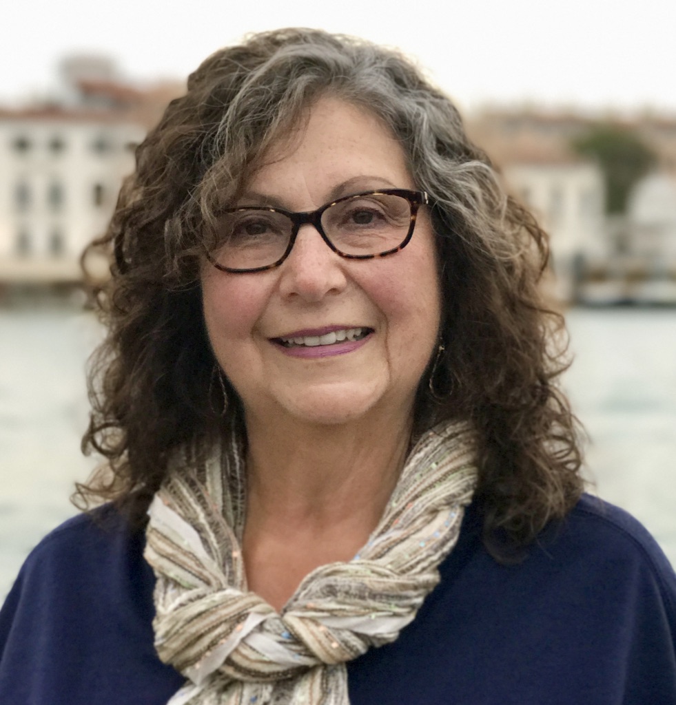 Smiling older woman with curly gray hair, glasses, and scarf, standing outdoors near a waterfront.