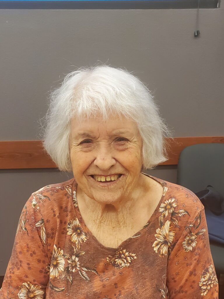 Smiling older woman with white hair wearing a floral shirt, seated at a senior services event.